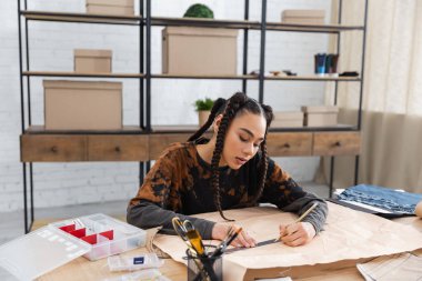 African american craftswoman drawing on sewing pattern near decor in workshop 