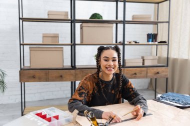 Smiling african american designer holding pencil near sewing pattern in workshop 