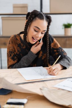 African american craftswoman drawing sketch and talking on smartphone in workshop 