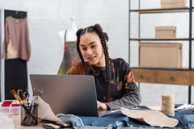 Smiling african american designer using laptop near cloth and sewing pattern in workshop 