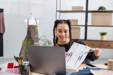 Young african american craftswoman pointing at sketch near laptop and sewing equipment in workshop 