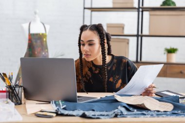 African american craftswoman holding sketch near laptop and cloth in workshop 