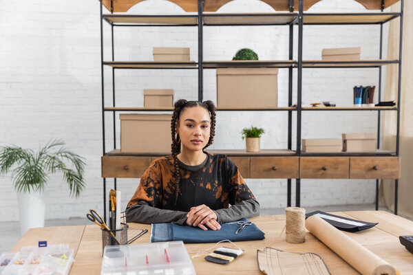 Young african american craftswoman looking at camera near cloth and sewing equipment in worktop 