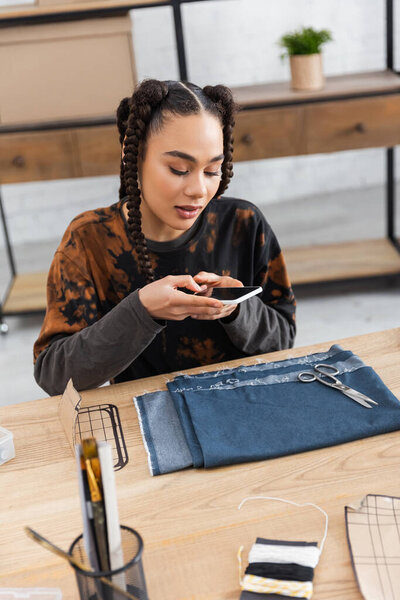 African american designer taking photo on smartphone near cloth in worktop 