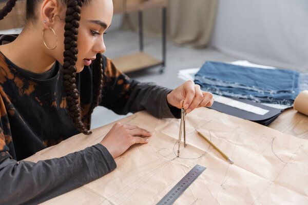 African american designer drawing with compass on sewing print near blurred cloth in workshop 