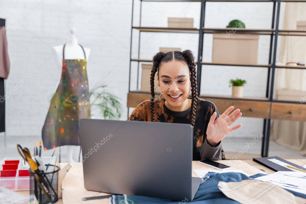 Smiling african american craftswoman having video call on laptop near smartphone and sewing equipment 