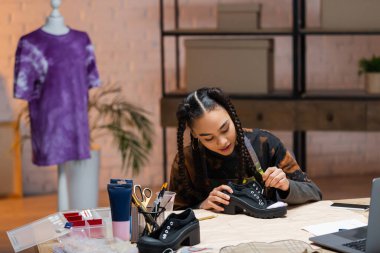African american designer painting on shoe near gadgets and sewing pattern 