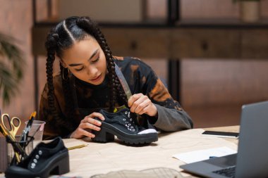 Young african american craftswoman painting on shoe near sewing patterns and devices in workshop 