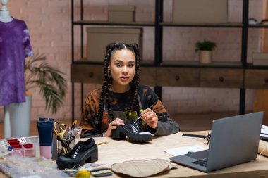 African american designer looking at camera while painting on shoes in workshop 