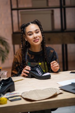 Positive african american designer holding paintbrush near shoes in workshop 