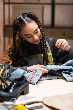 Young african american craftswoman embroidering denim jacket in workshop 
