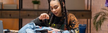 Young african american designer embroidering denim jacket in workshop, banner 