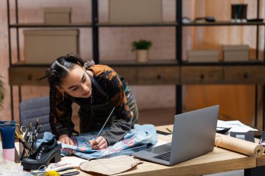 African american designer painting on denim jacket near laptop in workshop 