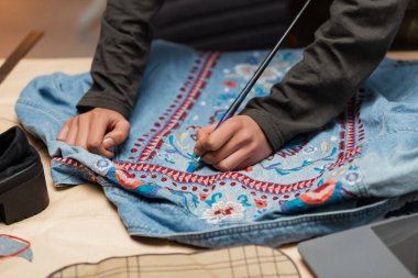 Cropped view of african american craftswoman painting on denim jacket near blurred laptop in workshop 