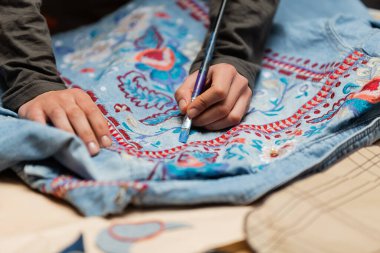 Cropped view of african american designer painting on denim jacket near blurred sewing pattern 