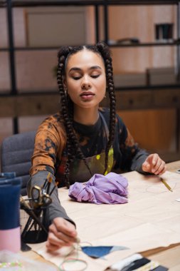 Young african american designer taking sewing equipment near cloth in workshop 