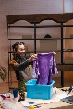Smiling african americn designer holding t-shirt near container in workshop 