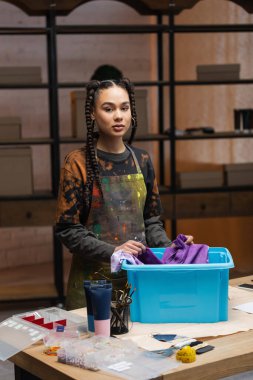 African american designer holding cloth near box and looking at camera in workshop 
