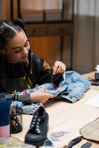 Smiling african american craftswoman embroidering denim jacket near sewing pattern in workshop 