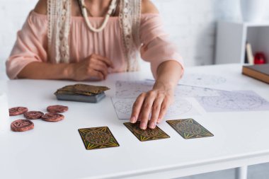 KYIV, UKRAINE - JUNE 29, 2022: cropped view of blurred astrologist taking tarot card near natal charts  and clay runes