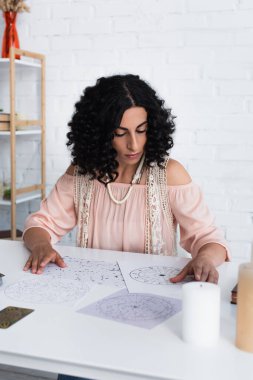 brunette astrologist near natal charts and candles on table