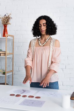brunette fortune teller with closed eyes standing near clay runes and cosmic charts during spiritual session