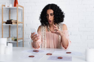 brunette soothsayer gesturing during video call on smartphone near clay runes and stars charts
