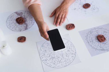 top view of cropped astrologist holding mobile phone with blank screen near celestial charts and clay runes
