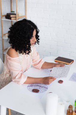 brunette astrologer looking at star charts near clay runes and prediction book clipart