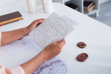 partial view of fortune teller holding cosmic charts near clay runes and prediction book