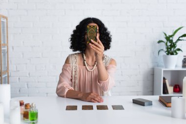 KYIV, UKRAINE - JUNE 29, 2022: brunette astrologer obscuring face with tarot card during spiritual session at home