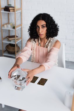 KYIV, UKRAINE - JUNE 29, 2022: brunette fortune teller looking at camera near crystal ball and tarot cards