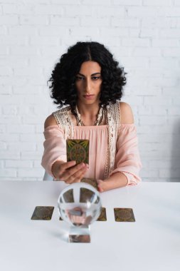 KYIV, UKRAINE - JUNE 29, 2022: brunette woman holding tarot cards near blurred crystal ball at home