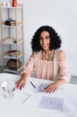brunette astrologer smiling at camera near crystal ball and star charts