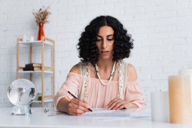 young astrologer drawing celestial map near crystal ball and candles