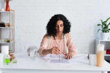 young astrologist drawing star charts near crystal ball and candles at home