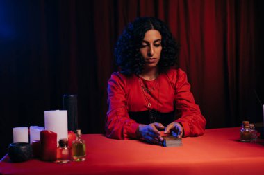brunette medium sitting near tarot cards and candles on dark background with red drape