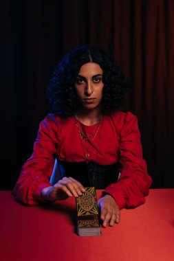 KYIV, UKRAINE - JUNE 29, 2022: young brunette predictor holding tarot card and looking at camera on dark background