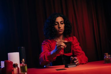 young fortune teller with tarot cards near candles and essential oils on dark background