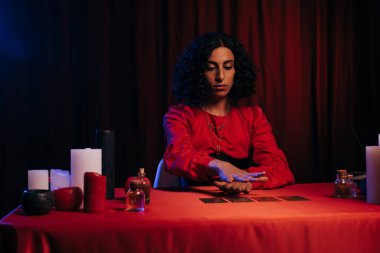 medium holding hand over tarot cards near candles and essential oils on dark background