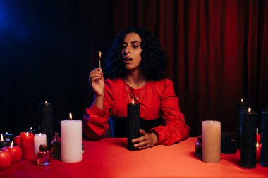 oracle with burning aroma stick near red table and candles in darkness