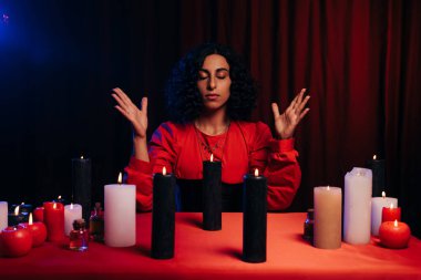 young oracle with closed eyes during spiritual session near burning candles on dark background with red drape clipart