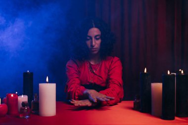 fortune teller sitting during magic session near burning candles on dark background with blue smoke 