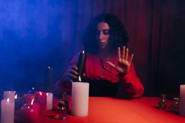 brunette oracle holding burning candle near red table on dark background with blue smoke