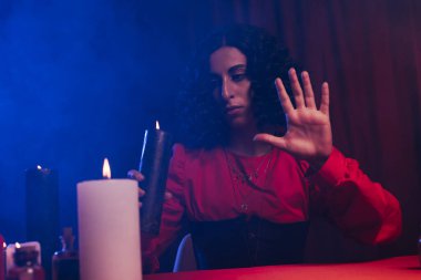 brunette fortune teller holding burning candle during spiritual session on dark background with blue smoke