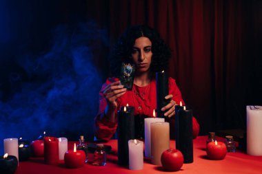 brunette fortune teller holding tarot card and burning candle on dark background with blue smoke