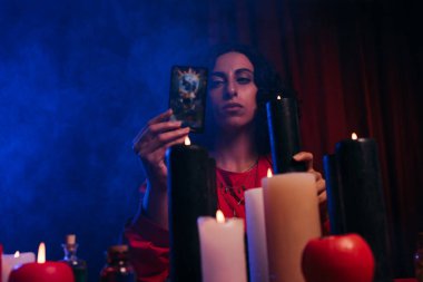 KYIV, UKRAINE - JUNE 29, 2022: young medium holding burning candle and tarot card on dark background with blue smoke