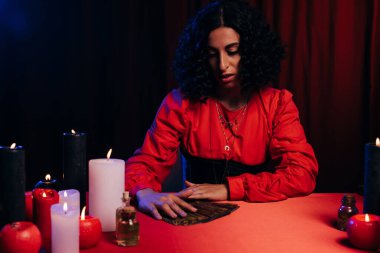 young brunette predictor near tarot cards and burning candles on red table and dark background