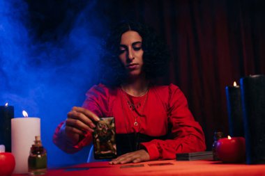 KYIV, UKRAINE - JUNE 29, 2022: brunette predictor holding tarot card near burning candles and blue smoke on dark background