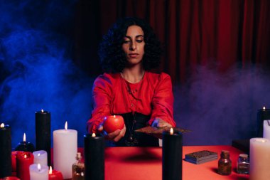 brunette fortune teller with candle and tarot cards near blue smoke on dark background